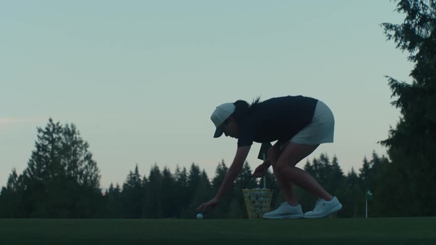 A female golfer walks onto the putting green and places balls from a basket to practice at twilight