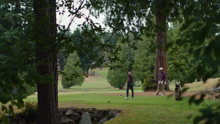 A man takes a powerful golf swing in slow motion on a lush green course as his friend watches on