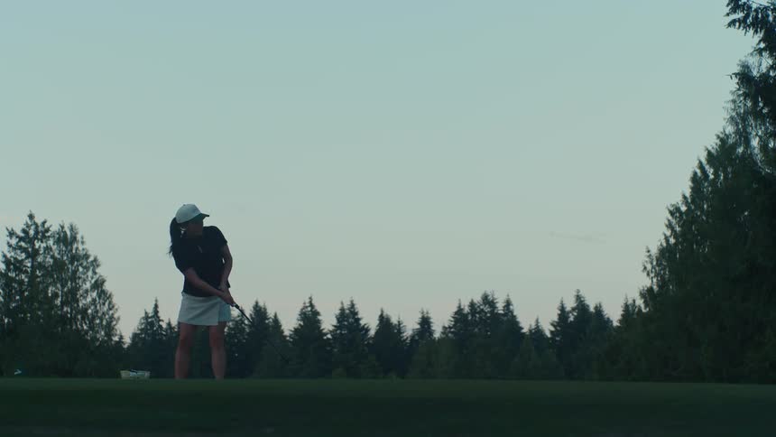 A female golfer practices her putting on a lush green course at dusk, with a dense forest in the background