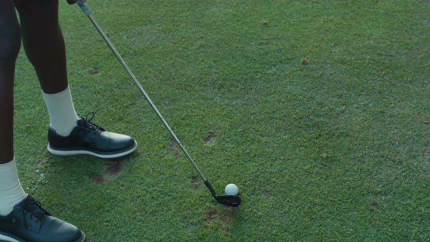 A slow-motion close-up reveals a golfer's legs and club hitting a ball before tilting up to their full swing on a sunny course
