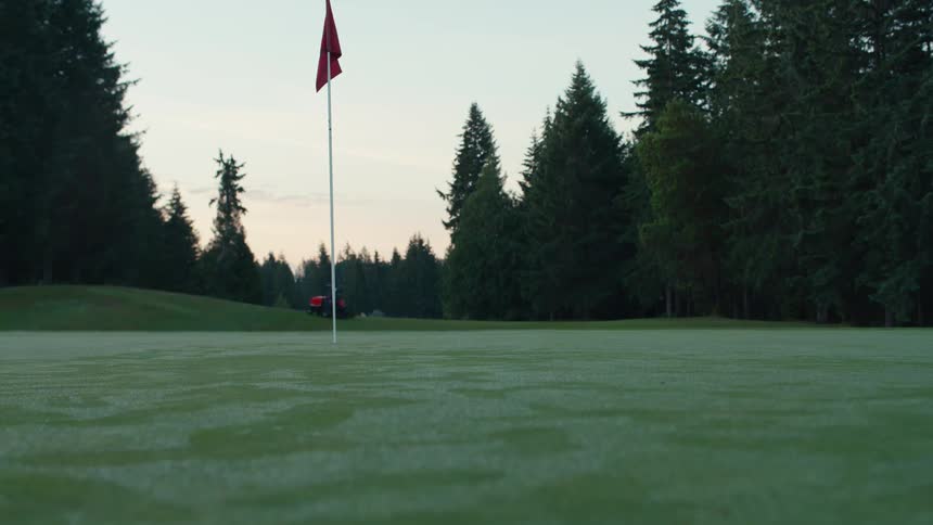 A groundskeeper on a riding mower cuts the dewy grass on a golf course putting green in the early morning