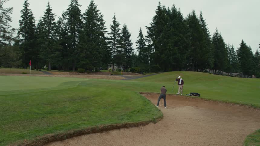 A man hits a golf ball out of a sand trap in slow motion while his friend watches from the green