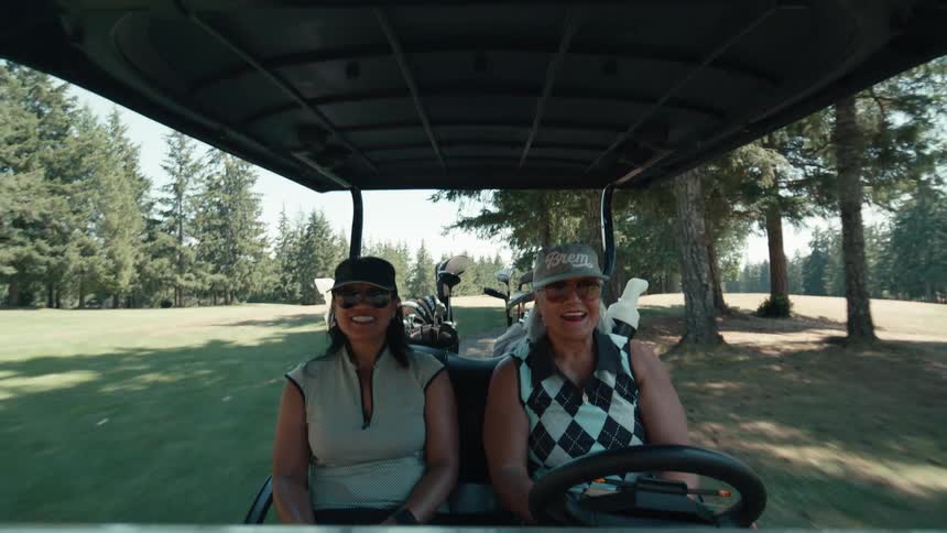 Dashboard point-of-view of two happy women driving a golf cart through a lush green course on a sunny day