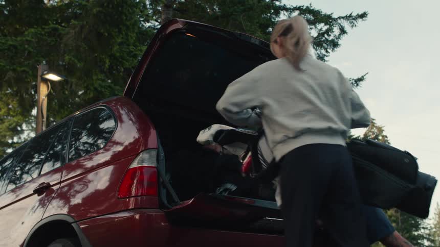 A man and woman unload their golf bags from the back of an SUV in a tree-lined parking lot
