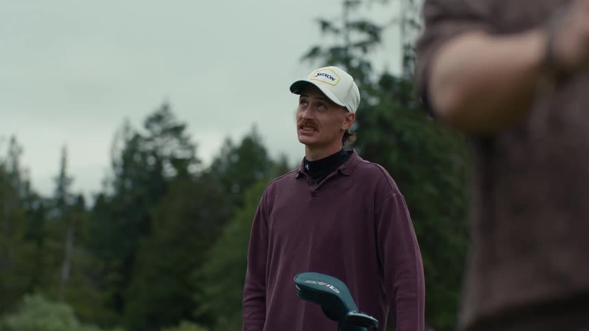 A young man with a mustache watches his friend swing a golf club in slow motion before offering some friendly advice on the course