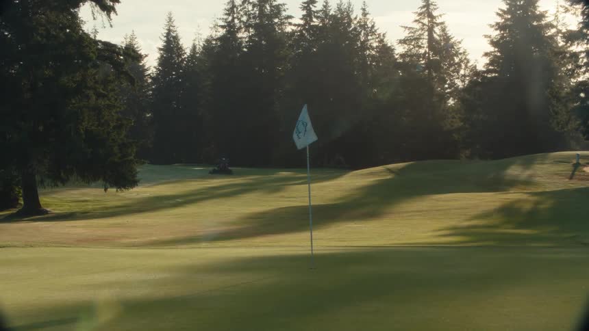 A peaceful golf course green is illuminated by the early morning sun as a groundskeeper mows the fairway in the background