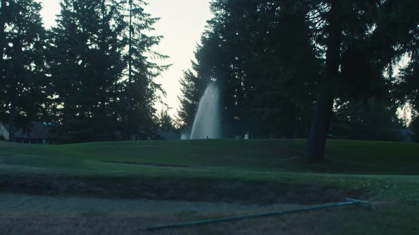 Lush green golf course is watered by sprinklers as the sun rises, with a golf cart driving by in the background