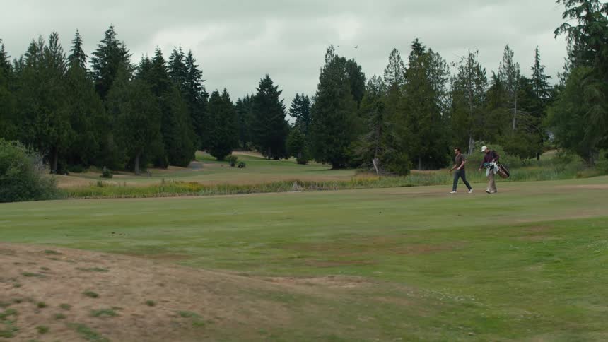Two men walk and talk while carrying their clubs across the lush green fairway of a golf course on an overcast day