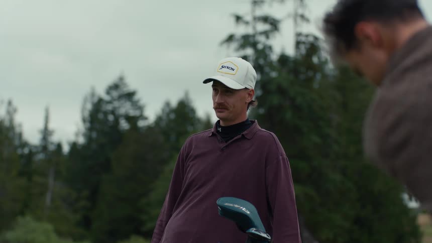 A man with a mustache watches and gives advice to his friend who takes a powerful golf swing in slow motion on the course
