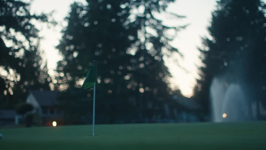 A golf ball is putted across the green at dusk, narrowly missing the hole as sprinklers water the course in the background