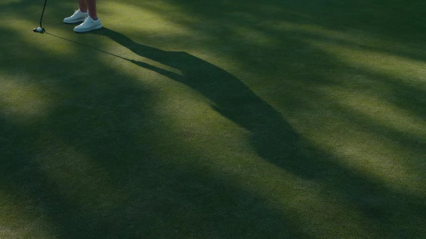 A golfer's long shadow is cast on the green as they putt the ball in slow motion, just missing the hole