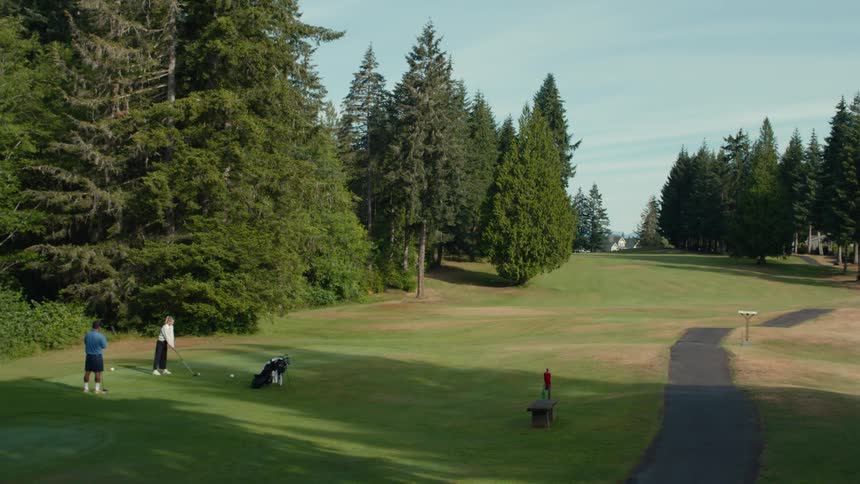 A female golfer tees off as her two companions watch from the tee box on a beautiful, lush golf course surrounded by tall evergreens