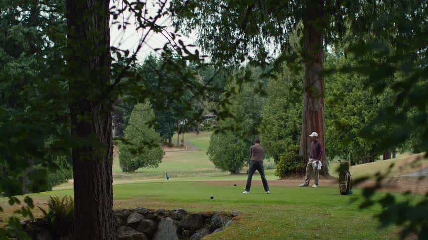 Framed by lush trees, a man takes a powerful swing with his driver on a beautiful golf course as his friend watches