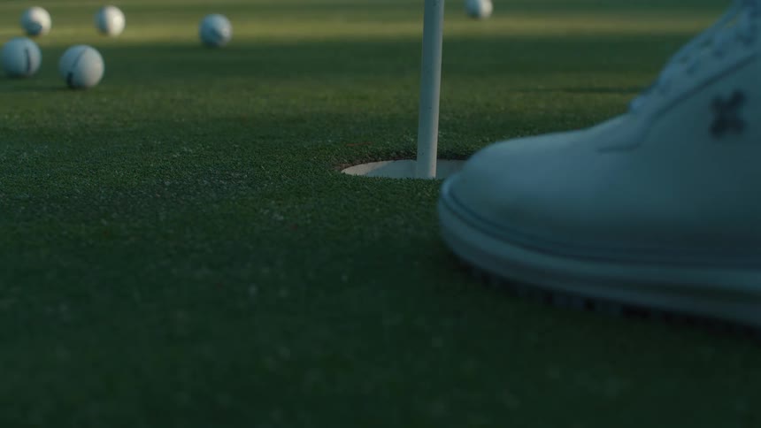 A golfer gathers scattered practice balls on a putting green, picking them up and dropping one into the hole