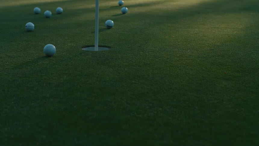 Slow-motion shot of multiple golf balls rolling across a putting green, with one successfully sinking into the hole