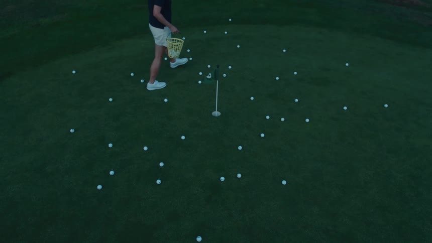 High-angle slow motion shot of a woman collecting dozens of golf balls into a basket after a putting practice session at dusk