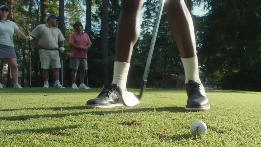 A golfer prepares to tee off, taking practice swings in a low-angle shot as his friends watch from behind on a sunny day at the golf course