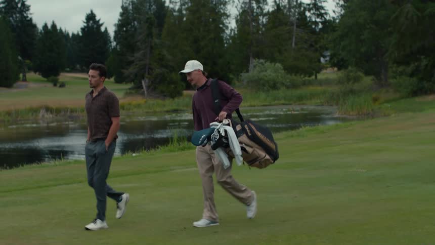 Two male friends walk and talk on a lush green golf course fairway, one carrying a golf bag