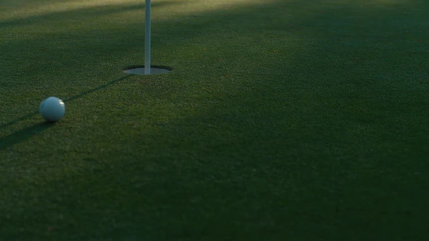 A golf ball putted in slow motion on a green misses the hole twice before sinking on the third attempt