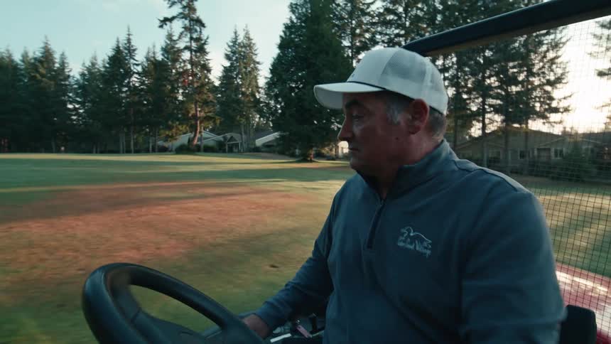 A golf course groundskeeper drives a red utility vehicle across the green fairway as the sun rises in the background