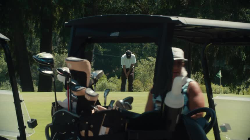 A man putts on a sunny golf course as the camera pulls back to reveal his friends getting into their carts