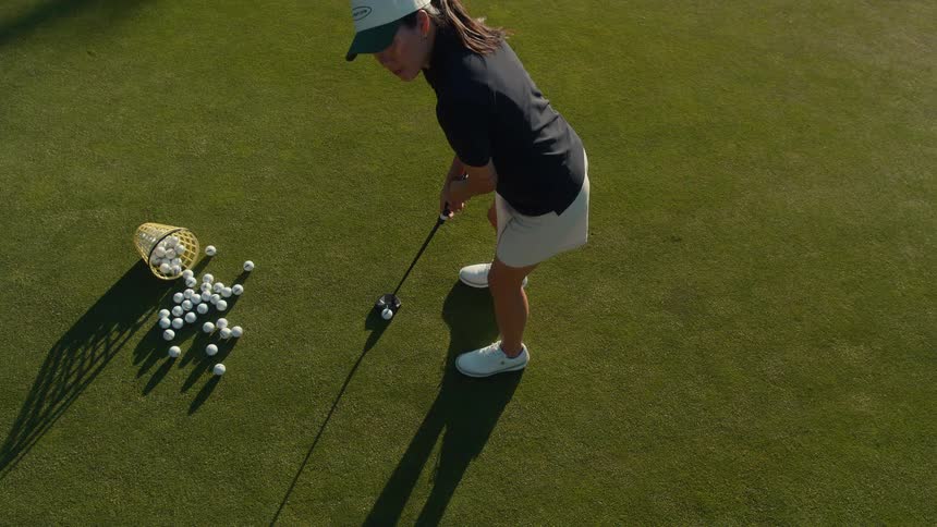 An overhead slow-motion shot shows a woman in a hat and golf attire practicing her putting on a pristine green course next to a basket of golf balls