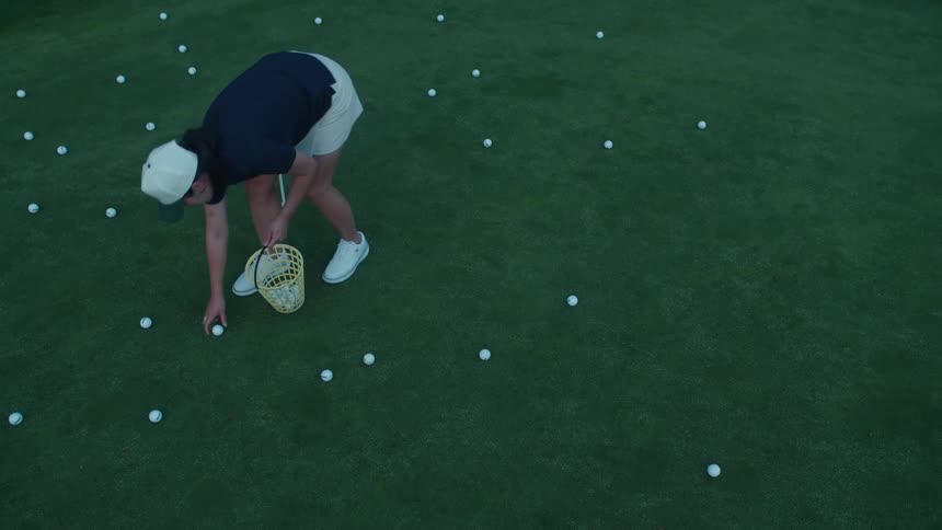 A golfer with a basket collects numerous practice golf balls from a putting green after a session