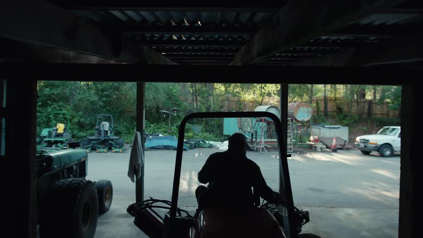 A man slowly reverses a red tractor into a dark, silhouetted garage filled with various farm equipment