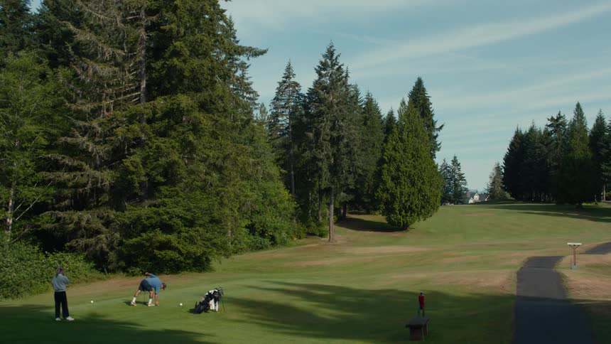 Friends tee off one by one on a beautiful, sunny day at a golf course surrounded by tall evergreen trees