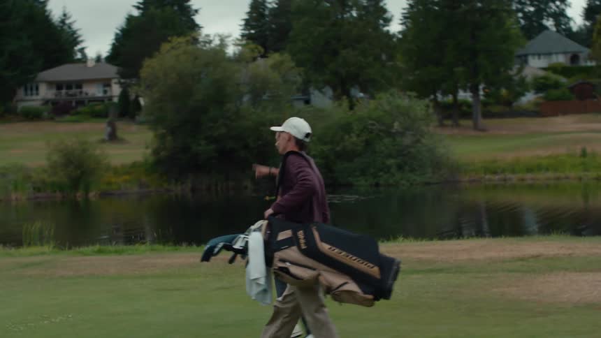 Two men walk across a beautiful golf course with a pond in the background, one carrying a bag of clubs