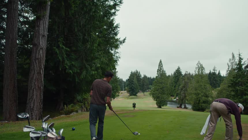 A man tees off in slow motion as his friend watches on a beautiful, tree-lined golf course