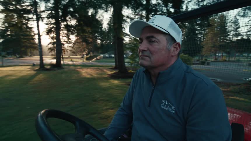 A groundskeeper drives a utility vehicle through a golf course as the sun rises in the background