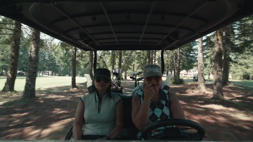 Point of view from the dashboard of two women talking and driving a golf cart through the trees on a sunny day