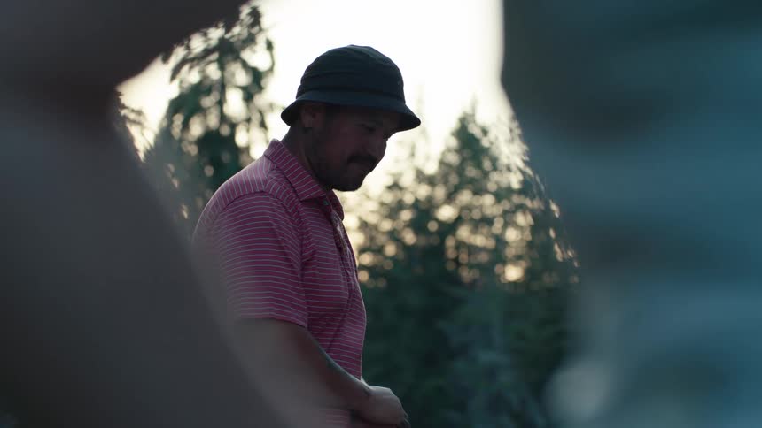 Framed between his friends, a man in a bucket hat and a pink polo shirt laughs and enjoys a conversation in a park at sunset
