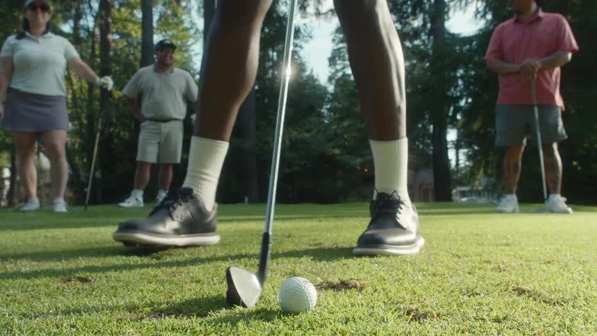 Slow-motion low-angle close-up of a golfer's feet and club as he sets the ball on the tee and swings while his friends watch