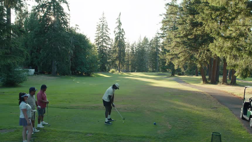 A man takes a powerful swing at the tee box in slow motion as his friends watch on a beautiful golf course