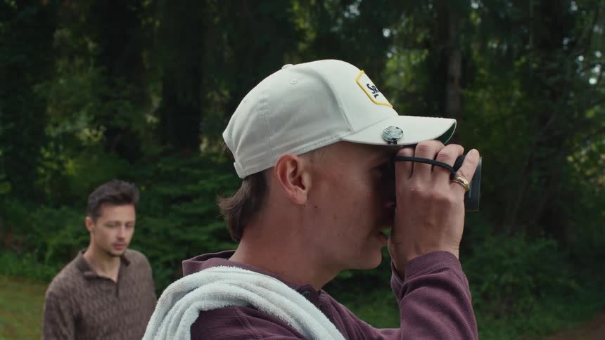 A man with a mustache and mullet uses a rangefinder on a golf course as his friend looks on