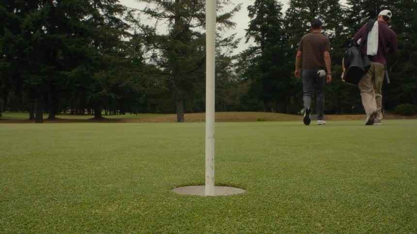 A golfer makes a successful putt in slow motion, retrieves the ball from the cup, and walks off the green with his playing partner