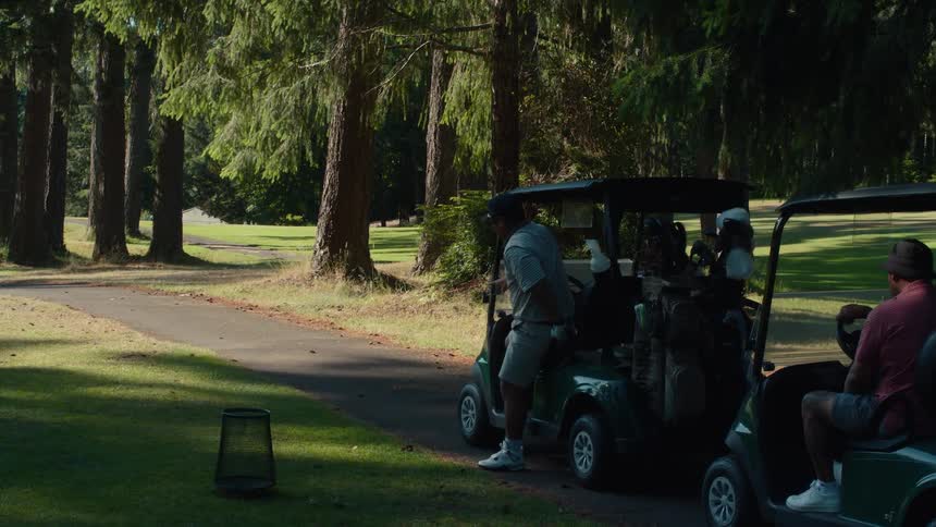 Golfers in carts drive along a tree-lined path before one prepares to tee off on a sunny day