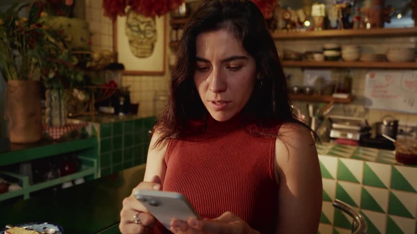 A woman sits at a quirky cafe counter and reacts with joyful surprise while looking at her smartphone