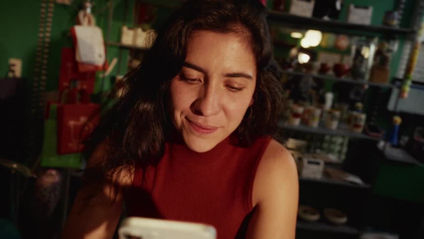 A woman smiles warmly while using her smartphone in a cozy, eclectic shop during golden hour