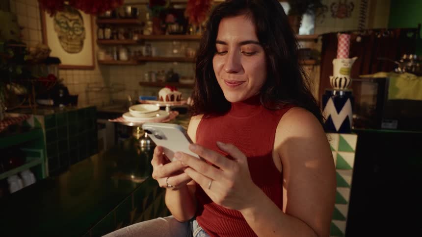 A woman sitting at a counter in a unique cafe smiles as she looks at her smartphone, illuminated by warm sunlight