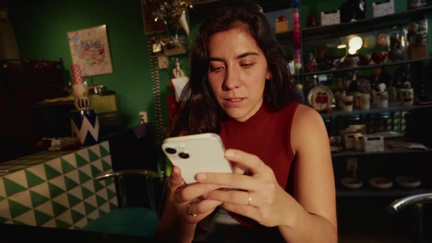 A woman sitting in a dimly lit, eclectic cafe smiles as she uses her smartphone