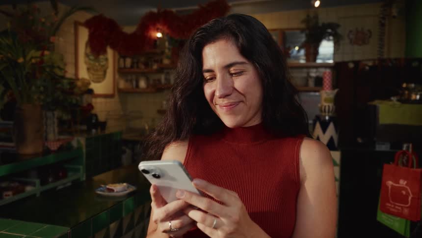 A lovely woman with dark hair smiles warmly as she reads a message on her smartphone while standing in a charming, uniquely decorated cafe