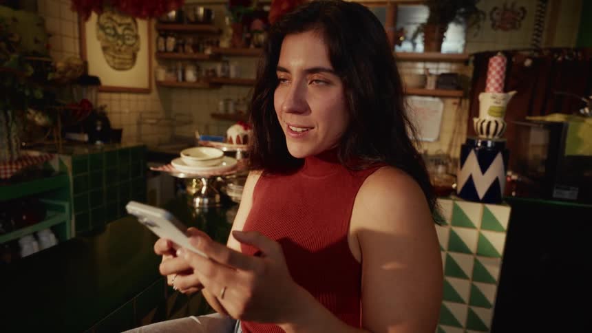 A woman sitting at the counter in an eclectic cafe smiles warmly as she looks up from her phone, bathed in sunlight