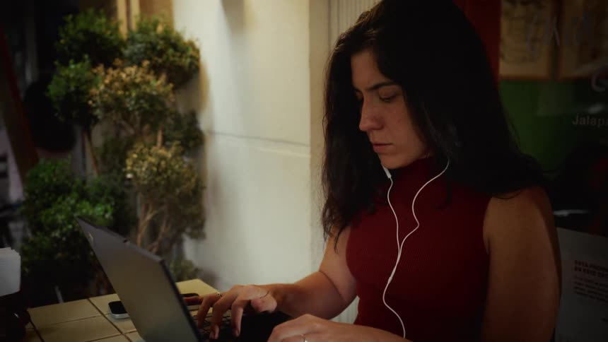 A focused young woman with earbuds works diligently on her laptop at an outdoor cafe at night