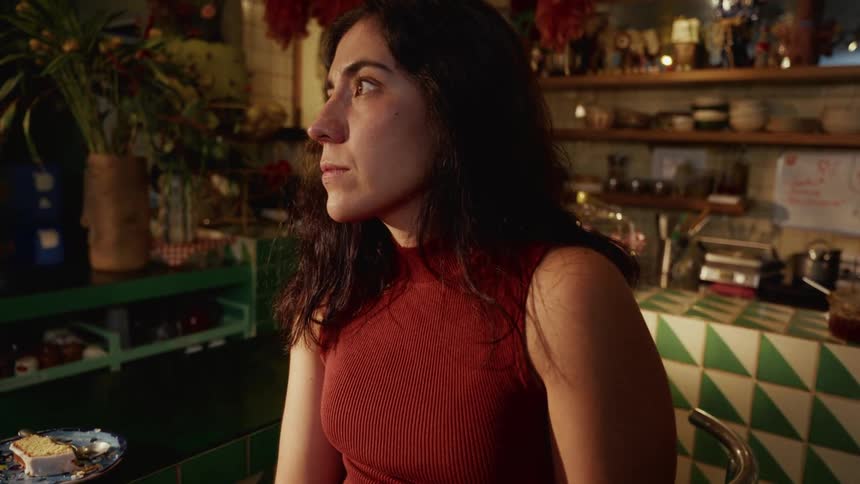 A woman scrolls on her phone at a cafe counter and then looks away with a serious and pensive expression