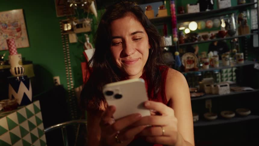 A young woman smiles warmly while looking at her smartphone in a cozy, eclectic shop