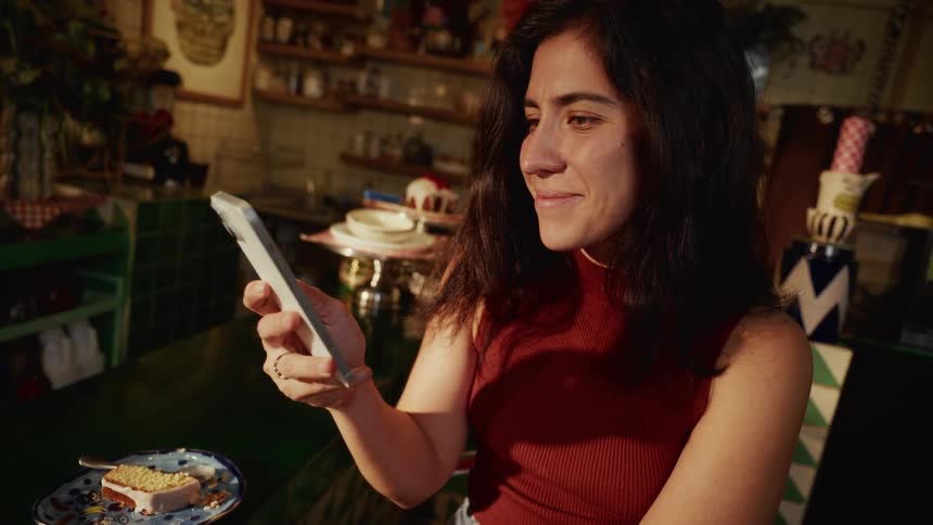 A young woman smiles warmly as she scrolls through her smartphone while sitting in a quirky, sunlit cafe