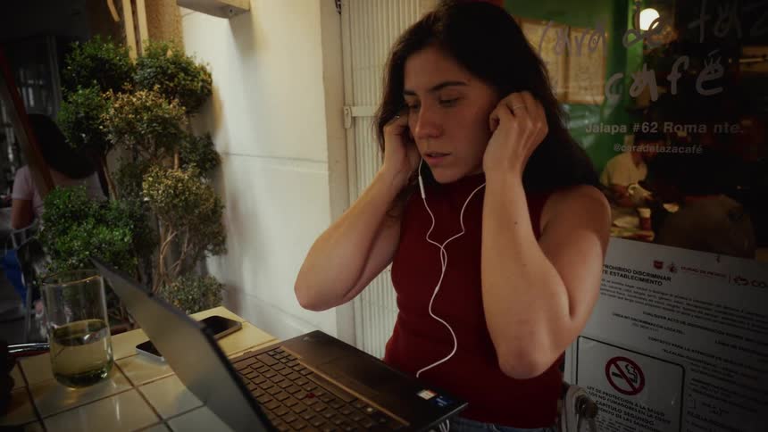 A focused young woman puts in her earbuds to work on her laptop at an outdoor cafe in the evening